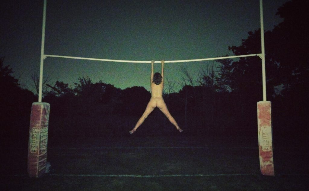 A naked woman hanging by her arms from a set of rugby goal posts. Photo. 