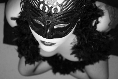 A woman wearing a mask and black lingerie kneels, looking up at the camera. Photo.