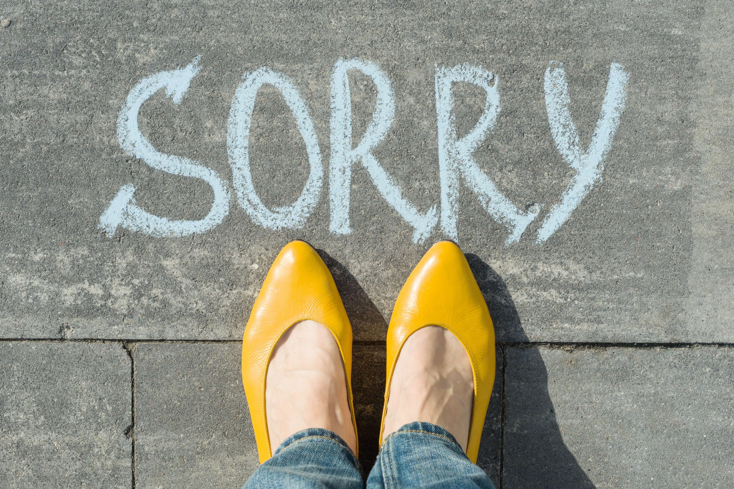 Femme feet wearing yellow heels next to 'sorry' written on asphalt in chalk. Photo.